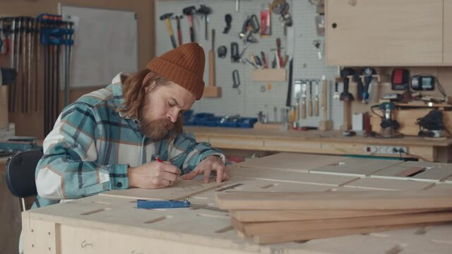 Lockdown of male Caucasian joiner with beard wearing casual clothes and hat sitting at workbench in workshop and making blueprint on piece of lumber