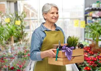 gardening, farming and old people concept - portrait of smiling senior woman in green apron holding wooden box with garden tools over greenhouse with flowers background