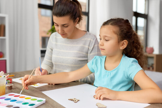 family, motherhood and leisure concept - mother spending time with her little daughter drawing or painting wooden chipboard items with colors at home