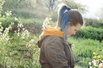 A teenage white girl with short blue hair is standing in the field surrounded by blooming trees. Backlit photo.