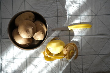 Potatoes in bowl, peeled potatoes, potato peels and knife on fabric.