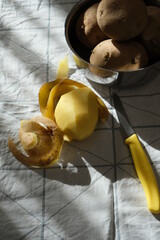 Potatoes in bowl, peeled potatoes, potato peels and knife on fabric.