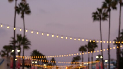 Defocused illuminated electric garland, palm trees silhouettes, Oceanside California USA. Ocean beach tropical pink sunset, pacific coast purple twilight sky. Los Angeles vibes. Bulb lights glowing.