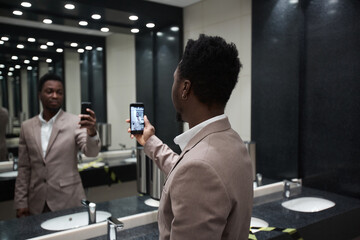 Portrait of African-American businessman taking mirror selfie in public restroom, copy space