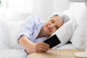old age and people concept - happy smiling senior woman with smartphone lying in bed at home bedroom