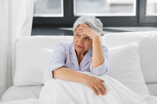 health, old age and people concept - senior woman in pajamas suffering from headache sitting in bed at home bedroom