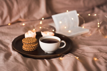 Cup of fresh black tea with stack of biscuits with chocolate filling on tray with burning candles and open paper book in bed over glow lights. Cozy home atmosphere.