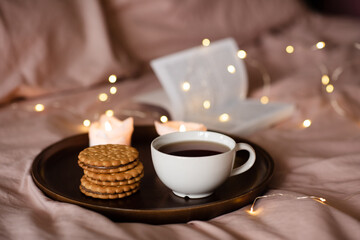 Cup of fresh black tea with stack of biscuits with chocolate filling on tray with burning candles and open paper book in bed over glow lights. Cozy home atmosphere.