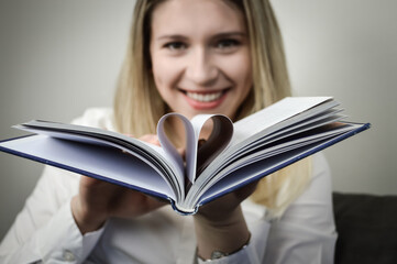 Portrait of a beautiful young blonde woman holding a book with the middle pages folded into a heart shape, concept for reading lovers