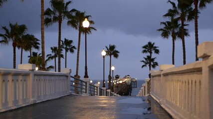 Rain drops, evening dark sky with clouds, Oceanside California USA. Empty pier and palm trees in twilight dusk. Reflection of lantern lights, illuminated wet broadwalk. Pacific ocean beach at night.