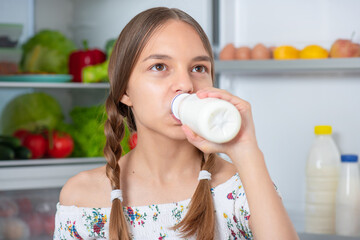 Beautiful young teen girl holding bottle of milk and drinks while standing near open fridge in kitchen at home