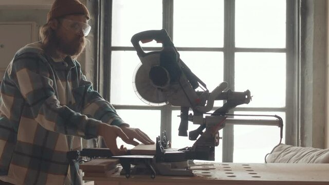 Medium shot of male Caucasian carpenter wearing checkered shirt, hat and goggles using automatic circular saw for cutting planks