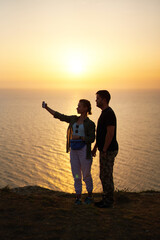 Fototapeta premium Couple is staying on the rock, taking photo and enjoying the sea view, sunset and the fresh air.