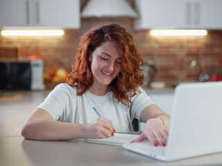 A beautiful young redheaded woman sits in the kitchen at the table with her laptop.