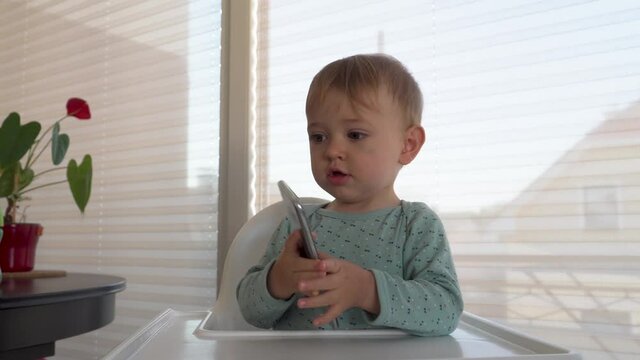 A cute baby talking to his mother on a cell phone in a baby chair. At home 4K