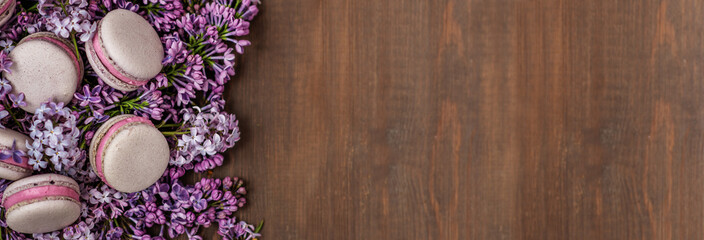 Obraz na płótnie Canvas Macaroni cakes lying on a bouquet of lilacs on a dark wooden background. Stretched panoramic image for banner