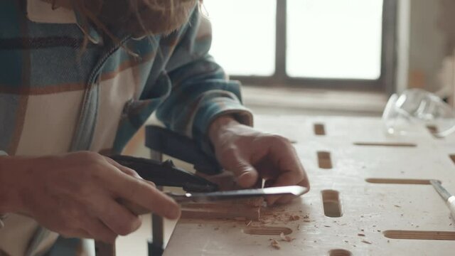 Close up of bearded male Caucasian carpenter wearing checkered shirt standing at workbench in joinery workshop and carving piece of timber with cutters