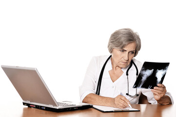 Serious elderly woman doctor sitting at table with computer and looking at x-ray