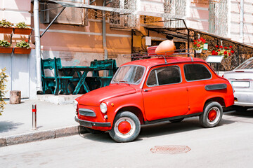 Red small retro car stand near restaurant as a decoration with old jug and leather luggage on the roof. Decorated. Attention. Street. Cafe. Sidewalk. Pavement. Outdoor. City