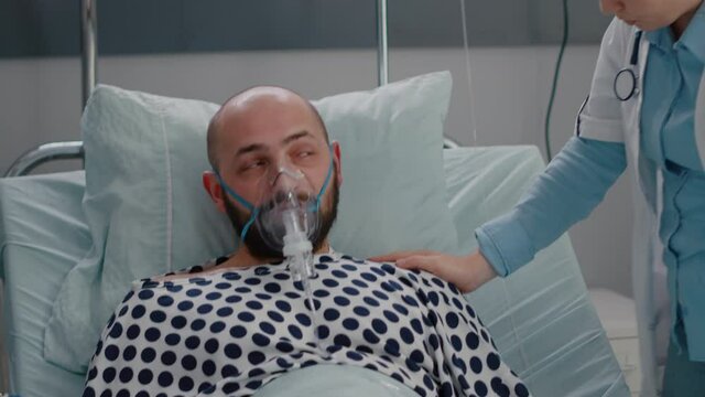 Close up of sick man patient resting in bed while doctor putting oxygen mask monitoring respiratory illness in hospital ward during healthcare emergency. Physician analyzing heartbeat pluse