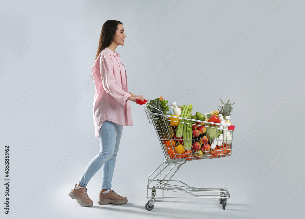 Poster Young woman with shopping cart full of groceries on grey background