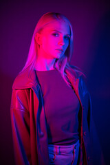 Fashion portrait of blonde woman in leather coat under neon light. Young stylish fashion model looks seriously into the camera.