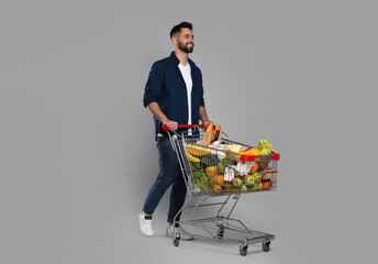 Happy man with shopping cart full of groceries on light grey background