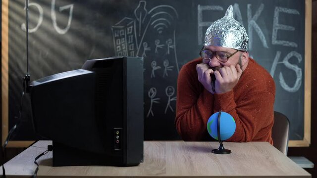 A bearded conspiracy theorist sits at his desk at home watching an old TV. A foil hat is put on the head. The paranoid is afraid. Conspiracy theory concept.