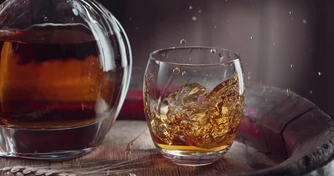 An ice cube slowly falls into a glass of whiskey on top of a vintage whiskey barrel. A decanter with a drink stands nearby, a dark brown background. 