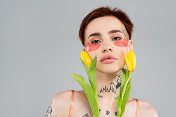 tattooed woman in pink eye patches holding yellow tulips isolated on grey