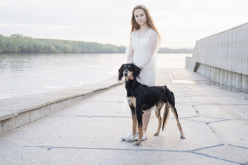 attractive young woman stand walk with saluki dog near river bank. 