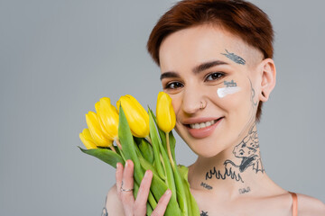 tattooed and happy woman with cosmetic cream on face holding yellow tulips isolated on grey