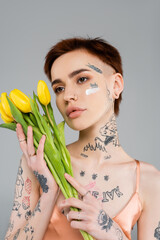 Naklejka premium tattooed young woman with cosmetic cream on face holding tulips isolated on grey