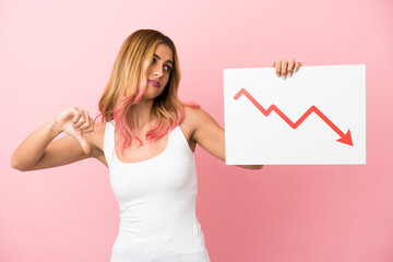 Young woman over isolated pink background holding a sign with a decreasing statistics arrow symbol and doing bad signal