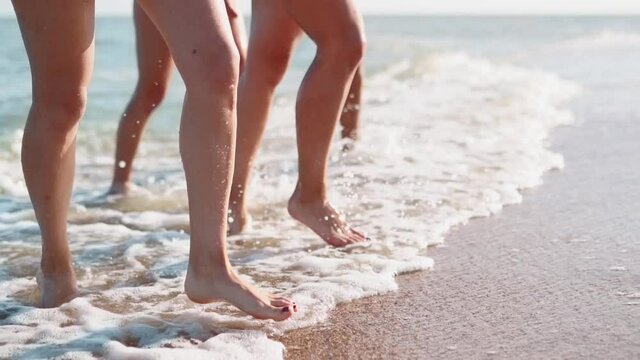 Slim female legs and feet walking out of sea water waves on sandy beach. Pretty women move on seafront. Splashes of water and foam in 120 fps slow motion. Girls after bathing in ocean go to the shore.