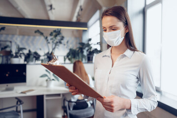 Fototapeta na wymiar Young businesswoman in face mask standing in office and holding clipboard with documents