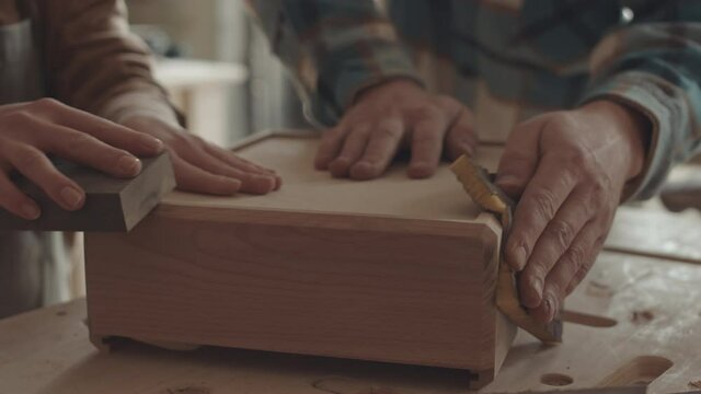Close up of hands of male and female joiners polishing wooden drawer lying on workbench using sandpaper