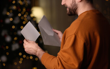 Man holding envelope and greeting card against blurred Christmas lights, closeup