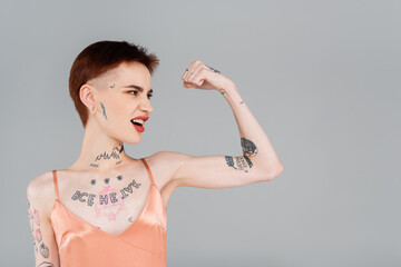 tattooed young woman with red lips showing muscle isolated on grey