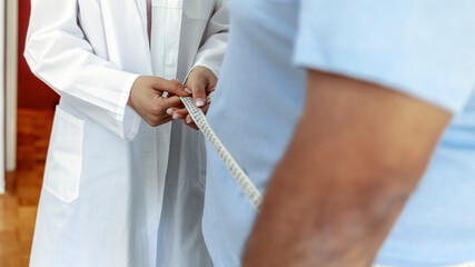 Overweight man with young female doctor in white coat standing in the medical room. Young woman doctor with tailors centimeter measuring overweight man. Unhealthy Lifestyle Concept.