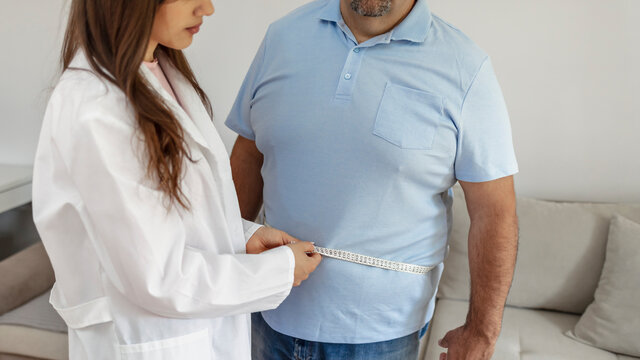 Cropped Shot Of A Female Doctor Taking Obese Patient's Body Fat Measurements. Dietitian Measuring Abdominal Circumference During Checkup. Young Doctor Taking Measurement From Depressed Overweight Man.