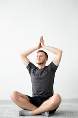 Yoga practice. Inspired man. Home meditation. Peaceful mind. Calm smiling guy in t-shirt shorts sitting lotus pose namaste hands up light wall interior background.