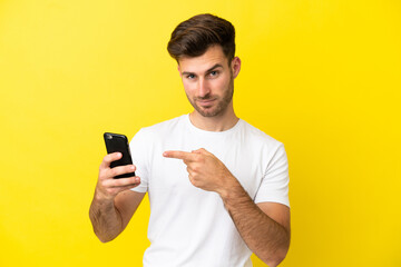 Young caucasian handsome man isolated on yellow background using mobile phone and pointing it