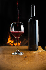 Wine, wine bottle and glass being filled with wine on rustic wooden surface with fire in the background, low key image, dark background, selective focus.