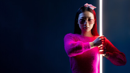 Cyberpunk fighter. Neon portrait. Laser war. Brave glamour Asian girl in pink in sunglasses in red light with glowing white LED lamp stick isolated on dark blue copy space background.