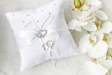 white pillow for wedding rings with rings and rhinestone hearts near white orchid on a white textured background. Wedding or engagement concept.