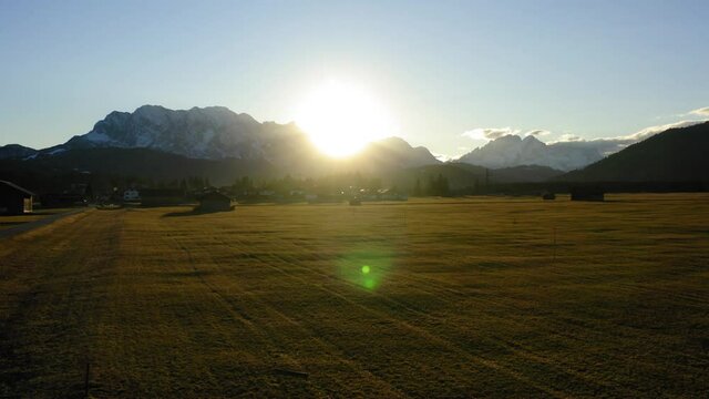 Drone shooting farmland with wooden houses located in valley in foot of snowy mountains, aerial bird's eye view of picturesque rural environment with homestead huts during sundawn