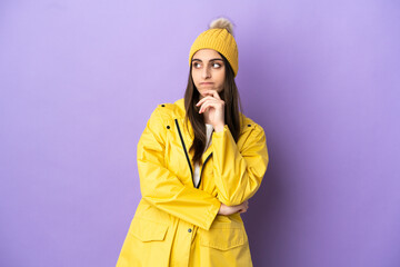 Young caucasian woman wearing a rainproof coat isolated on purple background having doubts