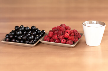 Ripe berries of raspberry and black currant on saucers on a wooden background. Slices or milk in a mug. Healthy breakfast in hot summer.