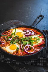 Shakshuka in an iron pan. Middle eastern traditional dish. Fried eggs with tomatoes, bell pepper, vegetables and herbs, sunny side up eggs. Vertical with copy space.